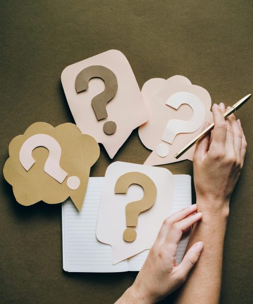 Hands holding pen next to paper question marks on notebook, symbolizing inquiry and creativity.