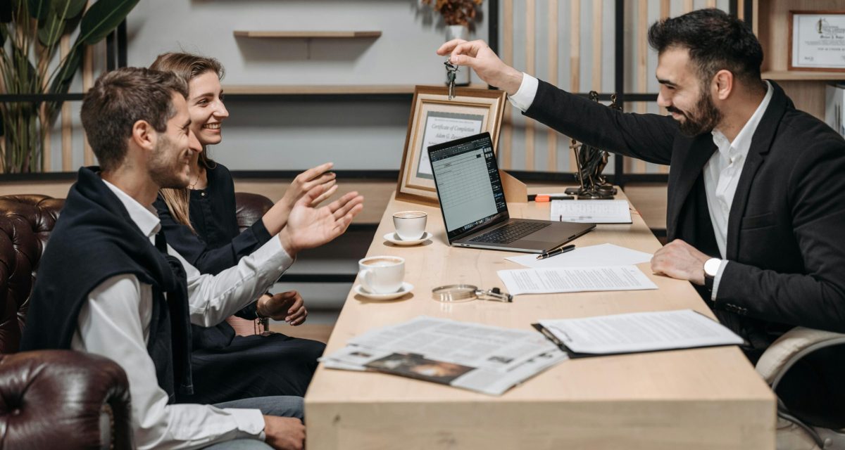 Business professional handing keys to smiling clients at an office desk during a consultation.